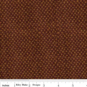 May include: A close-up of a brown fabric with a repeating pattern of small, gold stars. The fabric is textured and has a slightly fuzzy appearance. The fabric is measured in inches with a ruler at the bottom of the image. The ruler reads "1 Riley Blake 2 Designs 3 4 5 6".