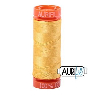 May include: A spool of yellow Aurifil thread. The thread is wound around a cylindrical core with orange caps at the top and bottom. The top cap has the brand name "AURIFIL" printed on it, and the bottom cap reads "100% CO".