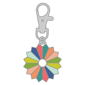 May include: A silver-toned metal keychain featuring a circular, multi-colored enamel charm. The charm is shaped like a stylized flower or sunburst with twelve distinct segments in shades of teal, orange, light pink, lime green, and navy blue.