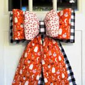 May include: A large, decorative bow with Halloween-themed fabric. The bow features orange fabric with white ghosts, pumpkins, and moons, paired with black and white buffalo check and white fabric with red polka dots. It is likely used as a door decoration.