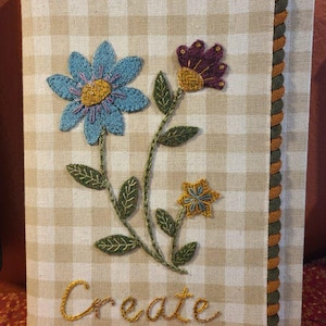 May include: A fabric-covered journal with a beige and white checkered pattern. Embroidered flowers in blue, purple, and yellow adorn the cover, along with the word "Create" in gold thread. A decorative braided trim runs along the spine.