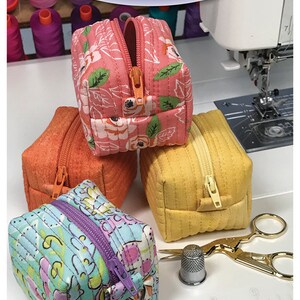 May include: Four small, square fabric zipper pouches in shades of orange, pink, yellow, and blue. The pouches are quilted and feature floral and geometric patterns. The text "Bodobo Bags" and "Bitsy Box" are visible.
