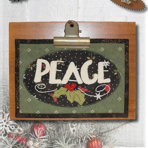 May include: A wooden clipboard holds a fabric mini-quilt that reads "PEACE" in white letters, with holly and berries. The quilt measures 5.5" x 8" and is on a green background. The skill level is easy.