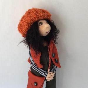 May include: A handmade doll with long curly black hair, wearing a red knitted beret, a striped long-sleeved shirt, a red knitted vest, green pants, and brown boots. The doll is carrying a red leather satchel.