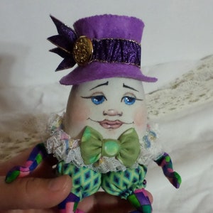 Puede incluir: Una muñeca caprichosa con forma de huevo con un sombrero morado, una pajarita verde y piernas de patchwork de colores. La muñeca tiene una cara pintada con ojos azules y una sonrisa.
