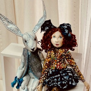 May include: A handmade doll with red curly hair wearing a colorful patterned dress and a blue bow sits next to a white rabbit doll wearing a gray vest. Both dolls are sitting on a white shelf.