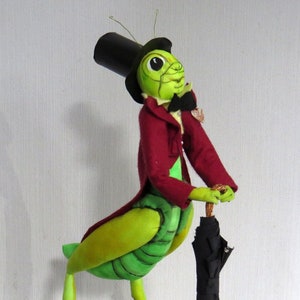 Può includere: Un grillo verde con un cappello a cilindro nero e una giacca rossa tiene un ombrello nero con un manico marrone. Il grillo è in piedi su una superficie grigia.