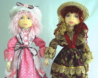May include: Two handmade dolls wearing vintage-inspired dresses and hats. The doll on the left has pink hair and a pink and white dress with lace trim. The doll on the right has red hair and a brown and gold dress with lace trim.