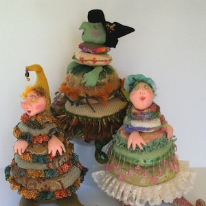 Może przedstawiać: Trzy ręcznie robione lalki z tkaniny o fantazyjnych rysach i kolorowych ubraniach. Lalki są ubrane w warstwy tkaniny i mają unikalne fryzury. Lalka po lewej stronie ma na sobie zieloną i złotą sukienkę z czapką z daszkiem. Lalka pośrodku ma na sobie zieloną i brązową sukienkę z czarnym kapeluszem. Lalka po prawej stronie ma na sobie zieloną i różową sukienkę z niebieskim kapeluszem.