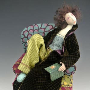 Puede incluir: Una muñeca hecha a mano con cabello castaño y un abrigo de terciopelo marrón está sentada en un sillón de colores. La muñeca sostiene un pequeño libro verde.