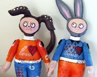 May include: Two whimsical handmade dolls, one with a bunny-like head and the other with a monkey-like head. The dolls are holding hands. One doll wears an orange shirt and blue pants, the other wears a blue shirt and orange pants. The shirts have hearts and text.