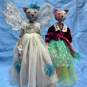 Op de afbeelding: Twee handgemaakte kattenpoppen die jurken en vleugels dragen. De pop links draagt een witte tule jurk met blauwe vleugels en een blauwe bloem op haar hoofd. De pop rechts draagt een paarse jas, een groene tule rok met witte stippen en een witte kraag.