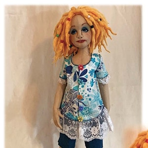 May include: A handmade doll called Ruth with long, curly, orange hair and blue eyes. She is wearing a blue and white floral dress with a lace trim, blue denim shorts, and blue sandals. The doll is standing on a white background. The text "Ruth, Pattern and Instructions, Diana Baumbauer Designs | Diana Baumbauer, Copyright 03.15.2021" is printed below the doll.