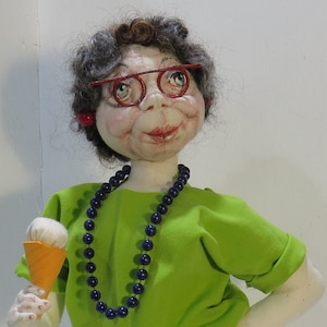 May include: A doll with gray hair wearing red glasses, a green shirt, blue pants, and a blue beaded necklace. The doll is holding a small ice cream cone.