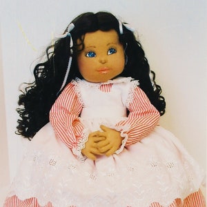 May include: A handmade doll with long dark brown curly hair, wearing a white and pink striped dress with a white lace trim and black shoes.