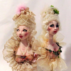May include: Two porcelain dolls with elaborate hairstyles and lace dresses. The dolls are on decorative stands. One doll has a pink feather in her hair and a pearl necklace. The other doll has green leaves in her hair and a green beaded necklace.