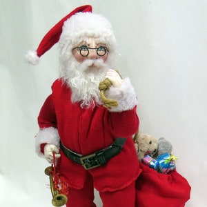 Könnte beinhalten: Eine Plüsch-Weihnachtsmann-Puppe, die einen roten Anzug mit weißen Verzierungen, einen weißen Bart und eine schwarze Brille trägt. Er hält ein Seil in der einen Hand und eine Spielzeug-Trompete in der anderen. Er steht neben einem roten Sack, der mit verpackten Geschenken und Plüschtieren gefüllt ist.