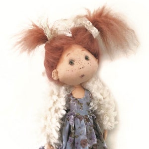 Pode incluir: Uma boneca feita à mão com cabelo ruivo em rabo de cavalo, vestindo um casaco de pele branco e um vestido azul floral. A boneca tem um rosto pintado com sardas e olhos castanhos.