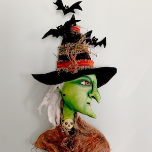 May include: A green witch doll with a long, pointed nose, wearing a black witch hat with a brown burlap brim and black bats. The doll has white hair and is wearing a brown burlap shawl with a skull pendant.