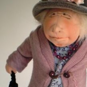 May include: A handmade doll with a pink and white flower crown, a gray hat, a pink cardigan, and a floral print dress. The doll is holding a black purse.