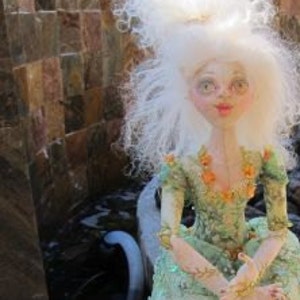 Könnte beinhalten: Eine Puppe mit weißen Haaren, die ein grün-weißes Kleid trägt und auf einem blau-weißen Sockel sitzt. Die Puppe trägt eine goldene Halskette und hat eine grün-goldene Handtasche.