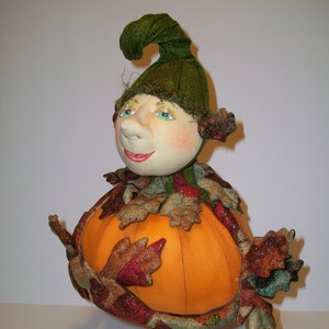 Puede incluir: Una muñeca de otoño caprichosa con un sombrero verde y un cuerpo de calabaza. La muñeca está decorada con hojas de otoño y tiene una cara amigable.
