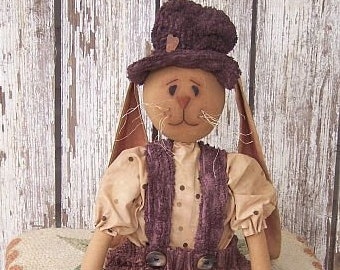 May include: A brown bunny doll wearing a brown hat, a brown and white polka dot shirt, and brown overalls. The doll is holding a small brown bag with green plants inside. The doll is sitting on a brown and white rug.