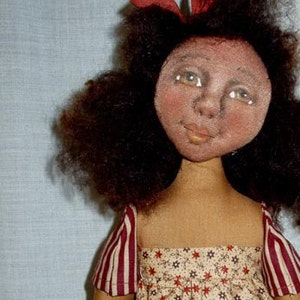 May include: A handmade rag doll with brown curly hair, wearing a red and white striped dress with a floral pattern. The doll is holding a smaller rag doll with brown hair and a white dress.