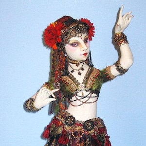 May include: A handcrafted doll wearing a red, gold, and green costume with a floral headpiece. The doll is posed with one arm raised and the other bent at the elbow. The doll is wearing a skirt with gold and green trim and a belt with tassels.