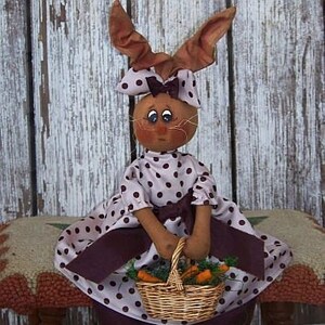 May include: A handmade fabric bunny doll wearing a pink and white polka dot dress with a brown sash and holding a basket of carrots. The doll is sitting on a wooden stool.