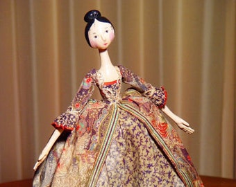 May include: A porcelain doll with black hair and a painted face wearing a long, patterned dress with gold and brown accents.