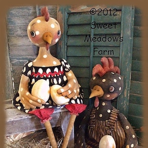 May include: Two handmade fabric chickens, one in a black and white polka dot dress and the other in brown and white overalls, each holding a white egg. The chickens are sitting on a bed of straw with a wooden bench behind them. The text "©2012 Sweet Meadows Farm" is in the upper right corner of the image.