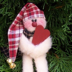 Könnte beinhalten: Eine handgemachte Schneemann-Puppe mit einem rot-weiß karierten Hut, einem weißen Pelzschal und roten Filzstiefeln. Der Schneemann hält ein rotes Herz in seinen Händen.