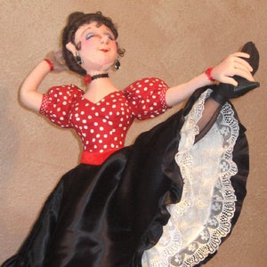 May include: A doll wearing a red and white polka dot dress with a black satin skirt and white lace trim. The doll is wearing black shoes and has her leg raised in the air.