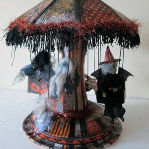 May include: A black, orange, and white Halloween carousel with a black fringe canopy, bats on top, and three figures: a witch, a ghost, and a black cat.