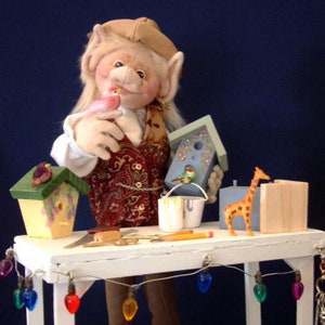 Könnte beinhalten: Eine fantasievolle Elfenpuppe mit blonden Haaren und einer roten Weste arbeitet an einem weißen Holztisch mit einer Lichterkette. Die Elfe hält ein blaues Vogelhaus und auf dem Tisch stehen weitere Vogelhäuser, ein Farbeimer, eine Giraffenfigur und ein Bleistift.