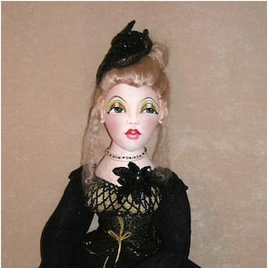 Puede incluir: Muñeca hecha a mano con cabello rubio, ojos azules y labios rojos. La muñeca lleva un vestido negro con detalles dorados, un sombrero negro y un collar de cuentas. Las manos de la muñeca están delicadamente posadas, y la estética general es vintage y elegante.