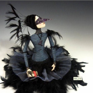 May include: A black and blue feathered doll with a long, black nose and a purple mask. The doll is wearing a blue and white striped dress and a black skirt. The doll is holding a small book in its hands.