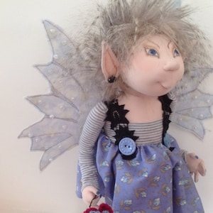 May include: A handmade doll with gray hair, blue eyes, and a blue and white striped shirt. The doll is wearing a blue and white floral skirt and has a pair of blue wings. The doll is holding a crocheted flower.