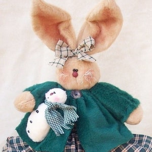 Könnte beinhalten: Eine Plüsch-Hasenpuppe, die eine grüne Jacke und einen karierten Rock trägt und eine weiße Schneemannpuppe hält. Der Hase hat braune Ohren und eine braune Schleife.