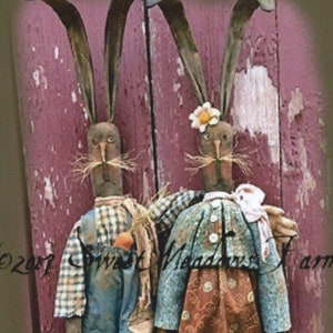 May include: Two handmade primitive bunny dolls with long ears. The bunny on the left is wearing a blue and white plaid shirt, blue overalls, and is holding carrots. The bunny on the right is wearing a brown and white floral dress with a blue jacket and a pink ribbon. Both bunnies have brown eyes and are standing on a wooden background.