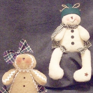 May include: A gingerbread person and a snowman doll with plaid accents. Both dolls have white trim and button eyes. The gingerbread person has a brown body and white trim around the edges. The snowman has a white body and a green hat.