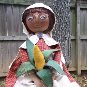 May include: A handmade rag doll dressed in a red and white checked dress with a white apron and a white bonnet. The doll is holding a green and yellow corn cob.