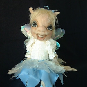 Könnte beinhalten: Eine fantasievolle Feenpuppe mit blonden Haaren, einem weißen Kleid und einem blauen Tüllrock. Sie hat schillernde Flügel und eine Libelle, die an ihrem Fuß befestigt ist.