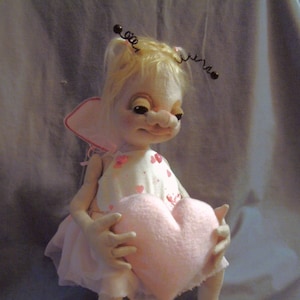 May include: A handmade doll with blonde hair, wearing a pink and white dress with hearts, holding a pink heart-shaped pillow. The doll is wearing black shoes with white lace trim.