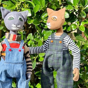 May include: Two handmade cat dolls wearing overalls and sneakers. One doll is gray with a striped shirt and red sneakers. The other doll is brown with a striped shirt and blue sneakers.