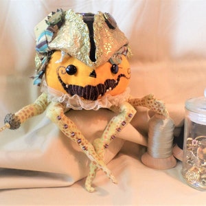 Puede incluir: Una muñeca de calabaza caprichosa con un sombrero de tela dorado y azul, un bigote negro y un cuello de encaje. La muñeca tiene piernas con manchas amarillas y marrones y está sentada sobre una superficie de tela beige. En el fondo hay una bobina de hilo y un frasco de botones.