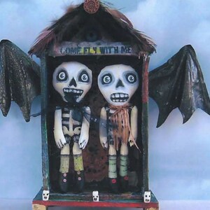 May include: Two skeleton dolls with wings stand in a small wooden house with the words "Come Fly With Me" painted on the front. The dolls are wearing colorful clothing and have painted faces.