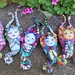 May include: Six colourful handmade fabric mermaid ornaments with beaded tails and whimsical faces. Each ornament features a different fabric pattern and colour scheme.