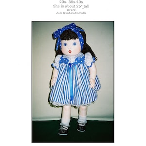 May include: A blue and white striped dress with a blue ribbon and white lace trim. The doll is wearing black shoes and has blue eyes, brown hair, and a red mouth. The doll is 26 inches tall.  "My Old Doll Beth" is the title of the image.  "Beth is a cloth reproduction of the old cloth and compo MA-MA dolls from the 20s- 30s-40s. She is about 26" tall. ©1979 Judi Ward-Judi's Dolls" is the text below the image.